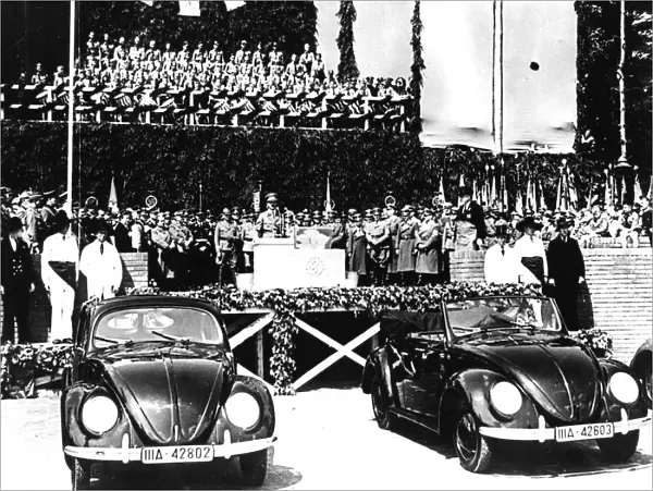 1930s Adolf Hitler: German Chancellor Adolf Hitler addresses a rally in the 1930s behind two Volkswagen Beetle cars